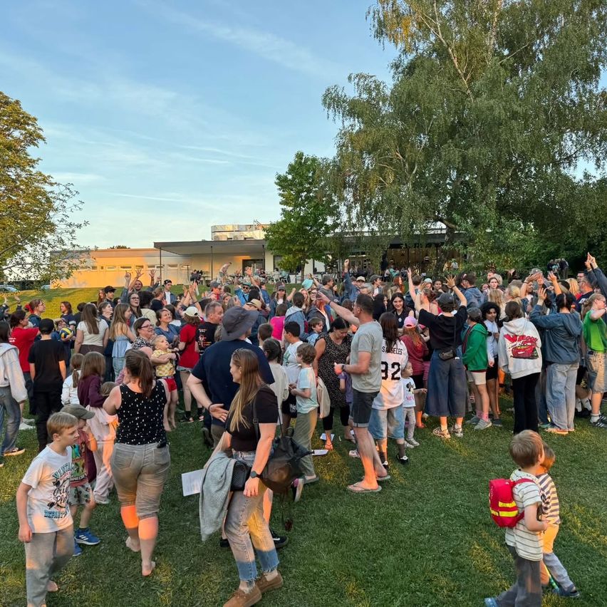 A large crowd of people gathers on a grassy field, some raising their hands. Trees and a building are in the background. Some individuals wear hats, glasses, and sneakers. The sky is clear with a few clouds.