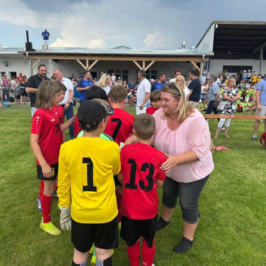 Eine Gruppe von Kindern in roten und gelben Trikots drängt sich auf einem Feld mit einer Frau. Zuschauer beobachten im Hintergrund.