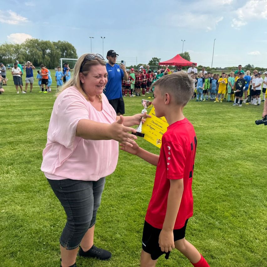 Eine Frau überreicht einem Jungen in einem roten Fußballtrikot einen Pokal. Sie stehen auf einem grünen Feld mit einer Menschenmenge im Hintergrund.