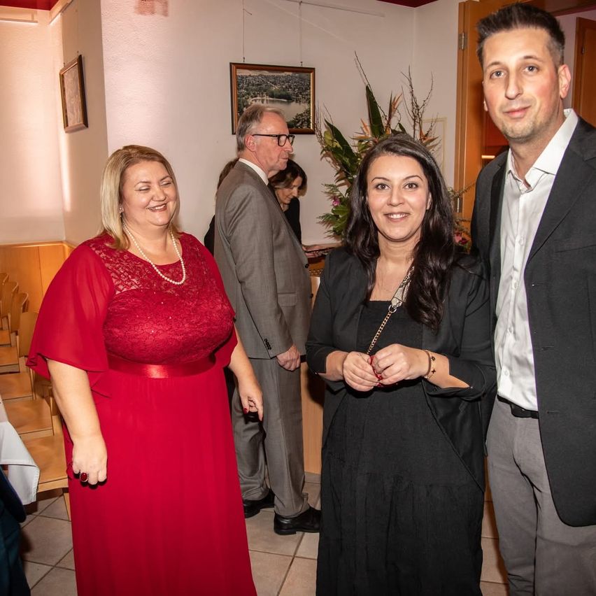 Ein Mann und zwei Frauen stehen in einem Raum zusammen. Die Frau auf der linken Seite trägt ein rotes Kleid, während die auf der rechten Seite ein schwarzes Kleid trägt. Der Mann trägt einen schwarzen Anzug. Hinter ihnen unterhalten sich ein weiterer Mann und eine Frau.