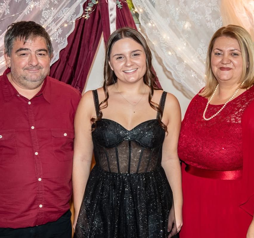 Ein Mann und eine Frau, beide lächelnd, stehen neben einer Frau in einem schwarzen funkelnden Kleid bei einer formellen Veranstaltung.