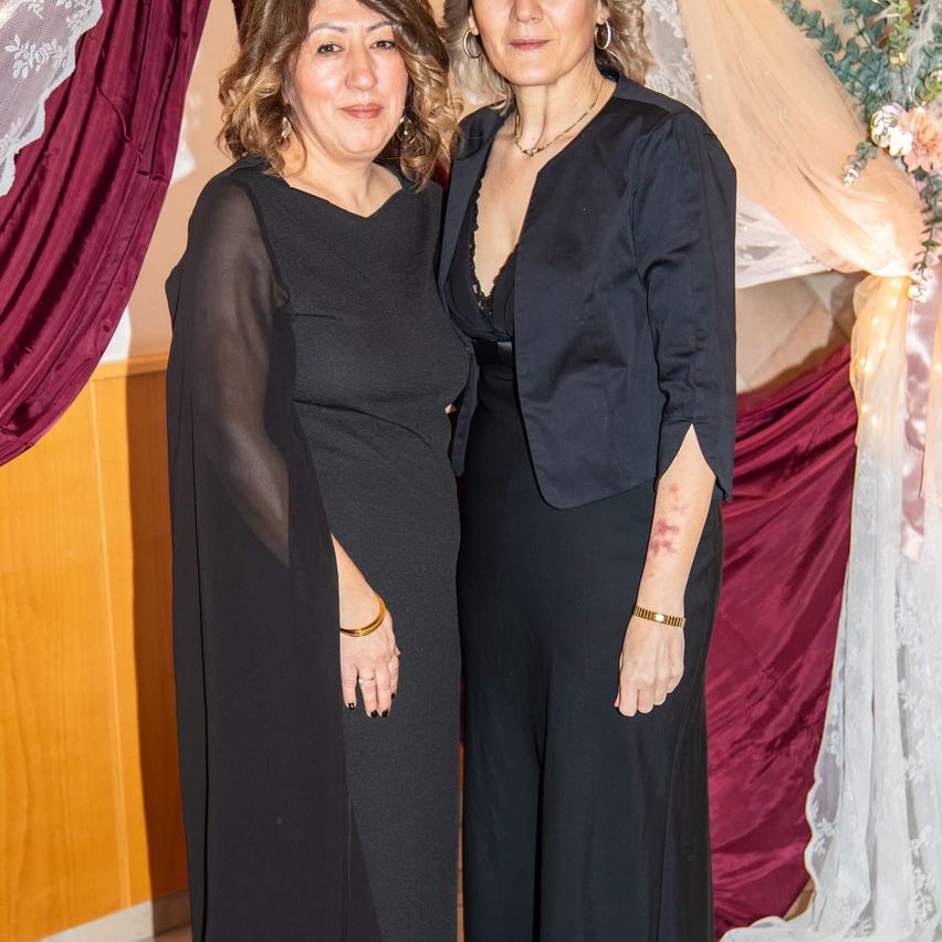 Zwei Frauen stehen nebeneinander und lächeln für ein Foto. Sie sind beide in Schwarz gekleidet. Die Frau auf der linken Seite hat ein goldenes Armband am linken Handgelenk. Hinter ihnen ist eine Kulisse mit einer Blumenarrangement.