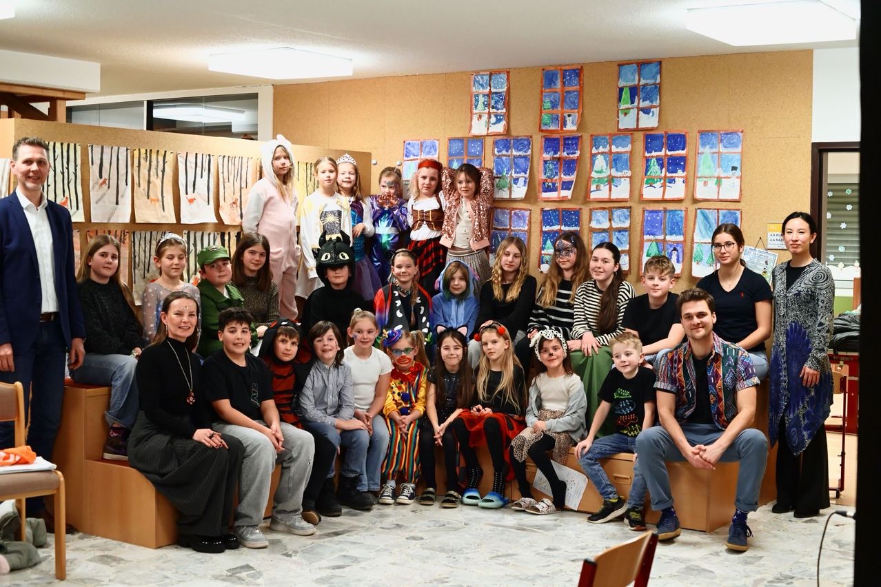 Eine Gruppe von Kindern und Erwachsenen posiert für ein Foto in einem Zimmer mit dekorierten Wänden, einige in Kostümen. Der Raum hat eine Lampe an der Decke und eine Wand mit gerahmten Bildern.