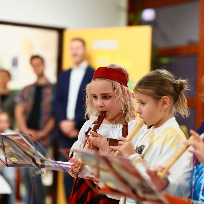 Zwei junge Mädchen in Kostümen spielen Flöten und stehen in einer Reihe in einem Klassenzimmer, mit verschwommenen Menschen im Hintergrund.