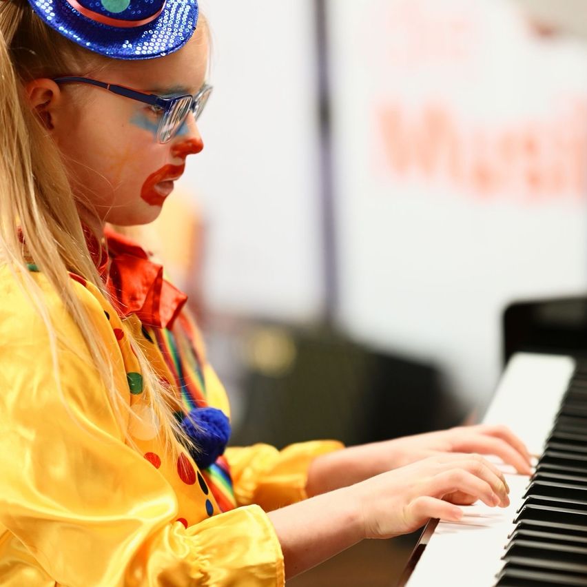 Ein junges Mädchen in Clownkostüm spielt mit beiden Händen Klavier. Ihr Gesicht ist rot und blau bemalt. Sie trägt eine Brille und einen Hut. Im Hintergrund ist ein Schild und ein verwischtes Bild.