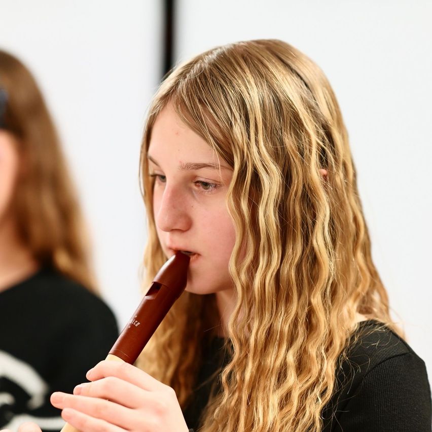 Ein junges Mädchen mit langen blonden Haaren spielt eine Holzblockflöte im Klassenzimmer. Sie trägt ein schwarzes T-Shirt. Eine weitere Mädchen mit braunen Haaren steht hinter ihr.