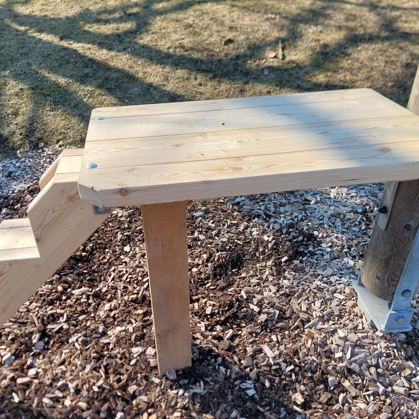 A wooden bench with a small staircase is placed on a playground, secured to the ground with metal brackets.