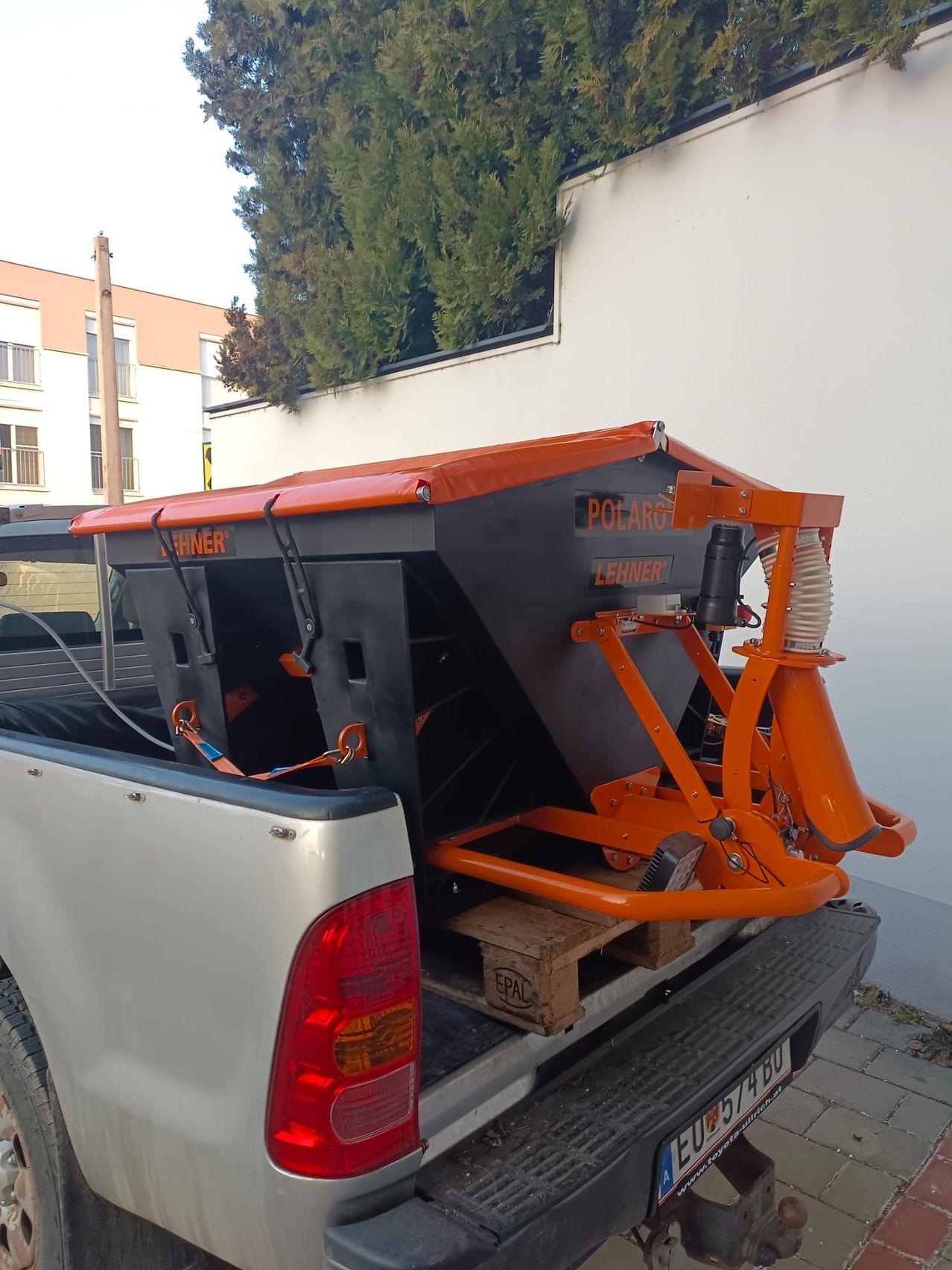 Ein orangefarbenes und schwarzes Gerät wird auf den Kofferraum eines silbernen Pickup-Trucks geladen. Die Maschine hat den Schriftzug Polargo an der Seite.
