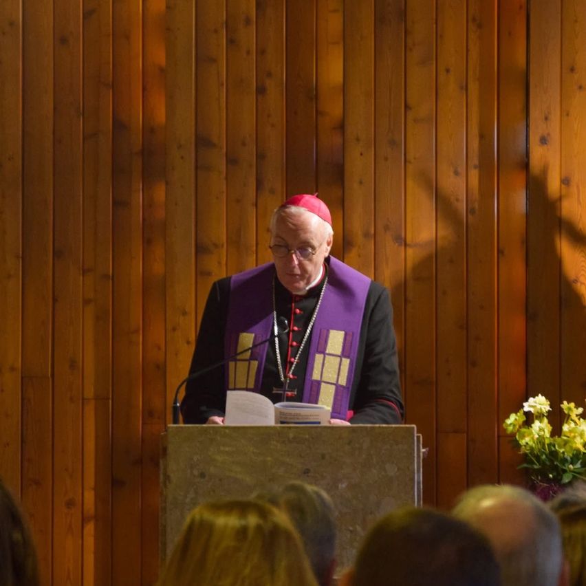 Ein Priester steht an einer Kanzel, trägt einen roten Hut und eine violette Stola und liest aus einem Buch. Das Publikum hört aufmerksam zu. Ein Strauß gelber Blumen ist rechts sichtbar.