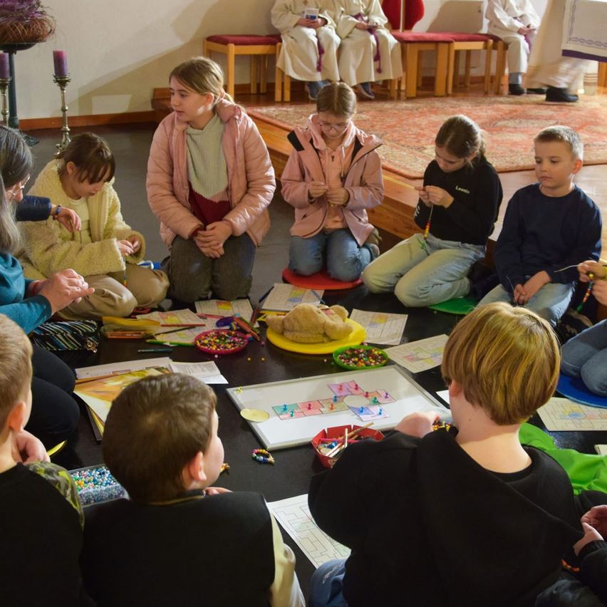 Kinder sitzen in einem Kreis um einen Tisch herum und sind mit einer Kunstaktivität beschäftigt. Einige tragen Jacken, andere tragen Hemden. Ein Stofftier und eine Kerze stehen auf dem Tisch. Zwei Erwachsene sind anwesend.