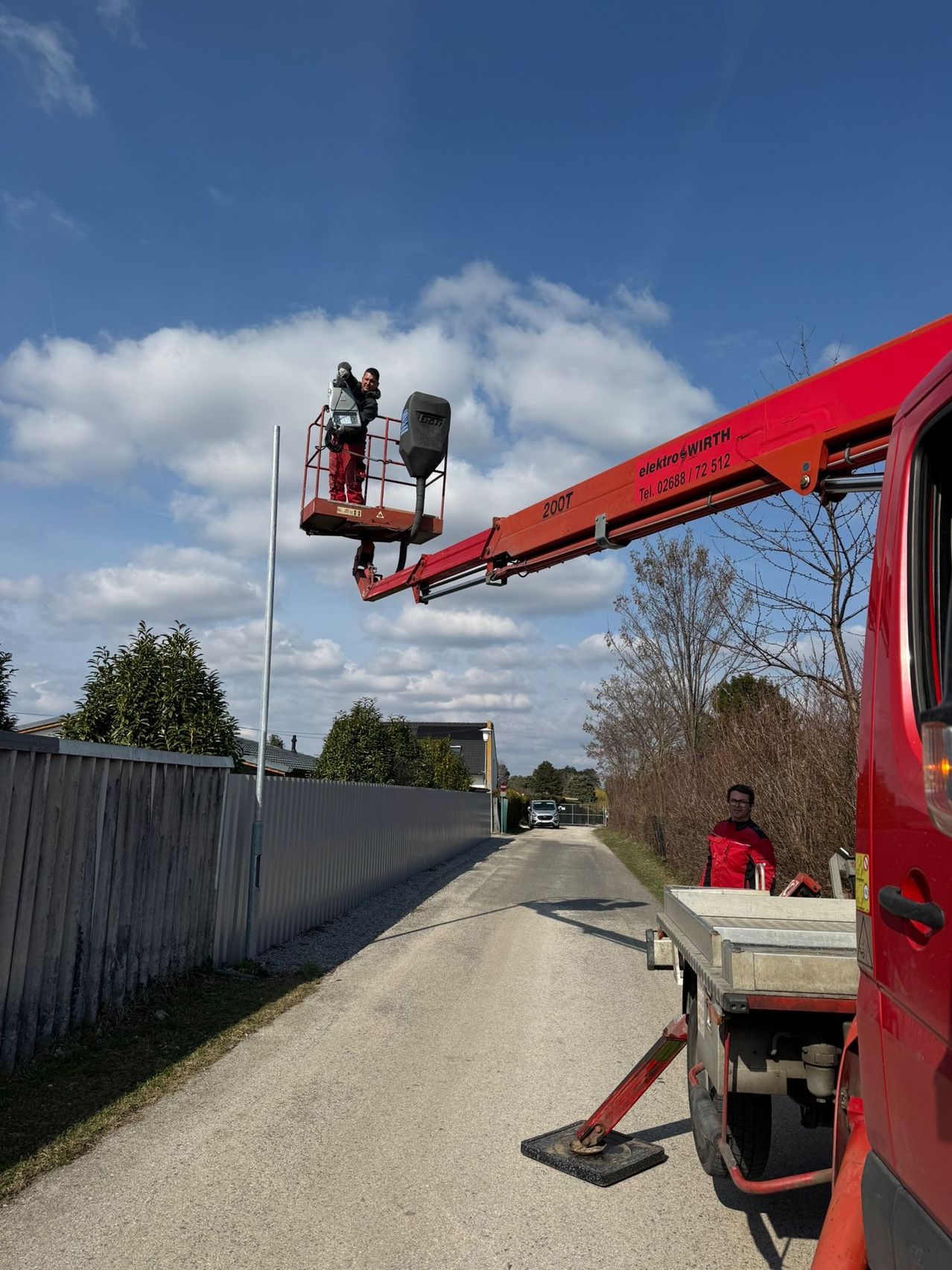 Ein Mann in einer Arbeitsbühne arbeitet an einem Versorgungsmast unter einem blauen Himmel mit Wolken. Ein anderer Mann steht in der Nähe, und ein Lastwagen ist am Straßenrand geparkt.