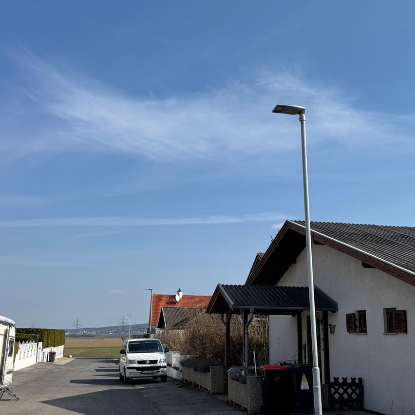 Eine Straße mit einem geparkten weißen Van unter einer Straßenlaterne, in der Nähe von Häusern, mit einem klaren blauen Himmel und einigen Wolken.