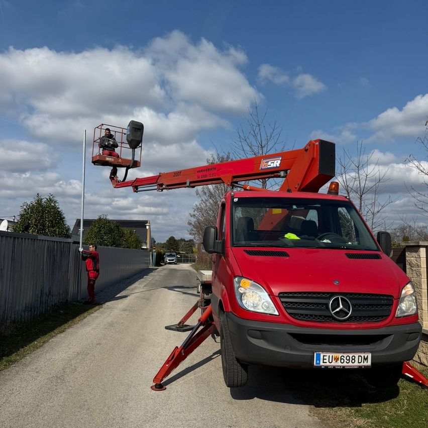 Ein roter Mercedes-Benz-Lkw mit einer Scherenarbeitsbühne ist auf einer Straße mit einem Zaun, einem Pfosten und einem Gebäude geparkt. Der Fahrer ist drinnen, und eine Person in einem roten Anzug steht in der Nähe des Zauns.