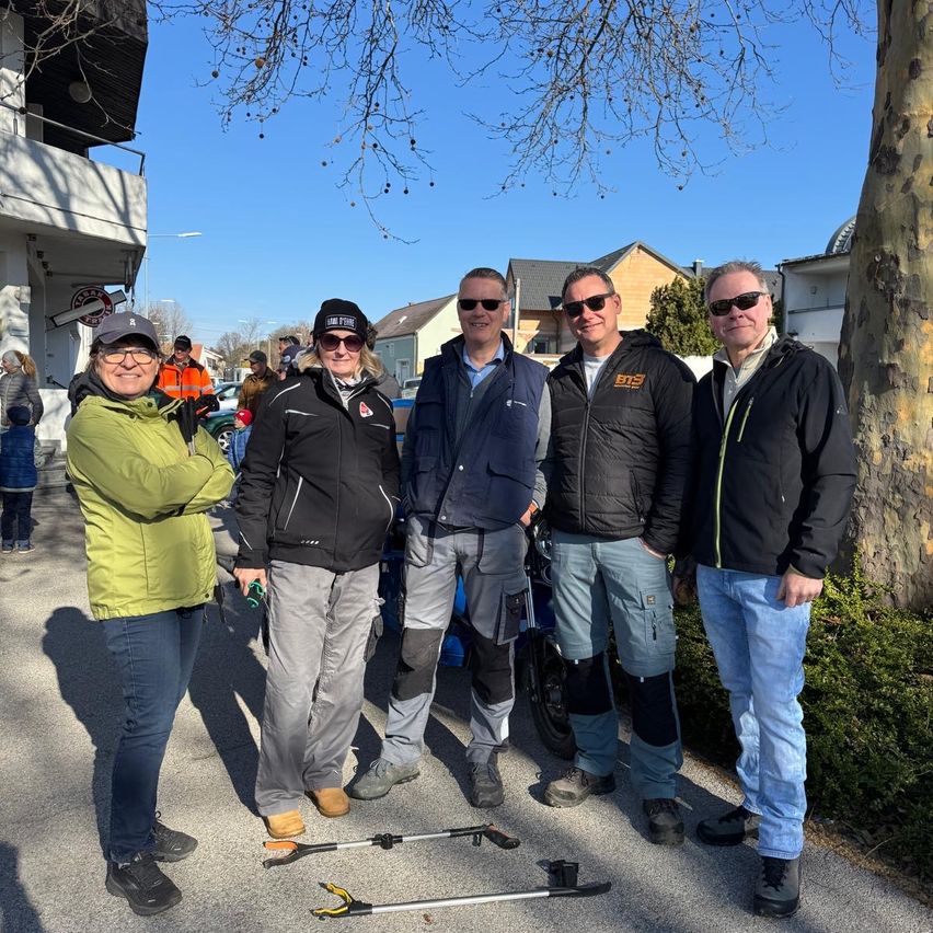 Fünf Personen stehen in einer Reihe und lächeln für ein Foto. Sie tragen Winterkleidung, wobei der Mann auf der rechten Seite ein Werkzeug hält. Hinter ihnen befinden sich Gebäude, ein Baum und mehrere Personen. Der Himmel ist klar.
