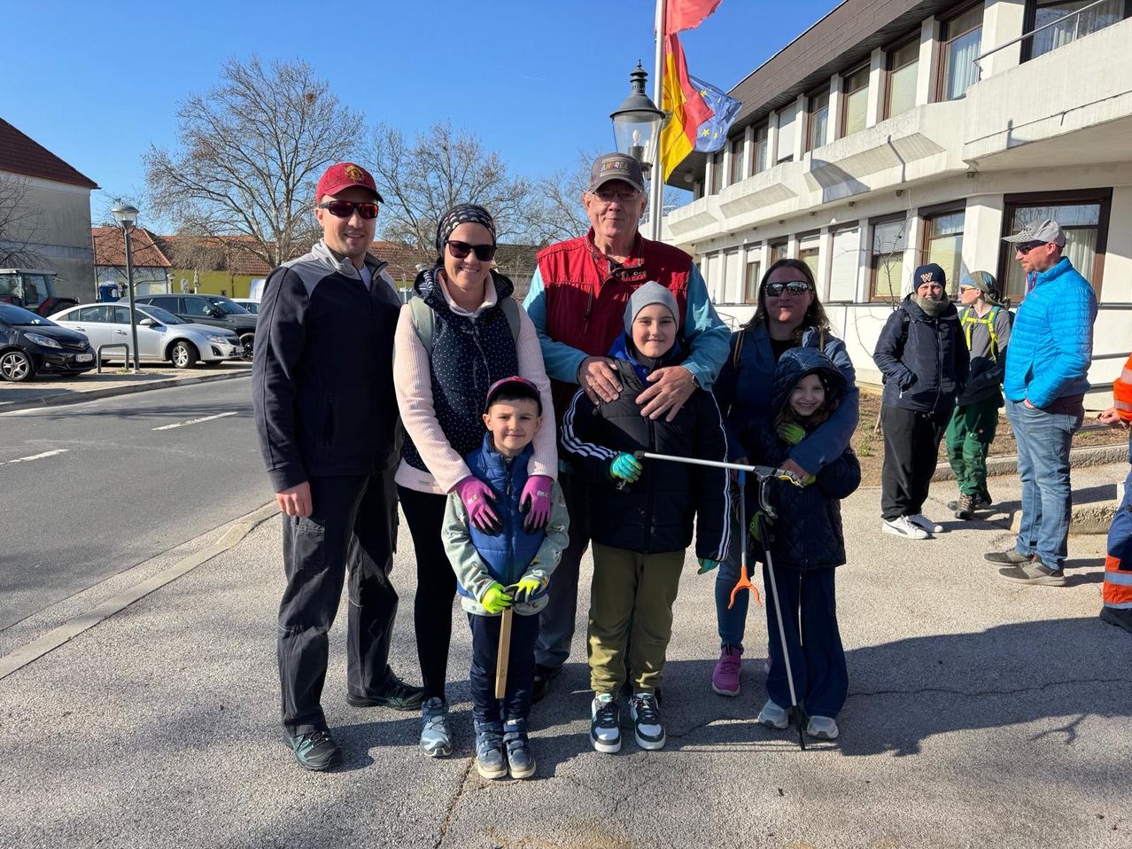 Eine Familie von sieben Personen steht an einem sonnigen Tag im Freien und lächelt für die Kamera. Sie halten Stöcke in den Händen und tragen Handschuhe. Ein Gebäude und Bäume sind im Hintergrund zu sehen.