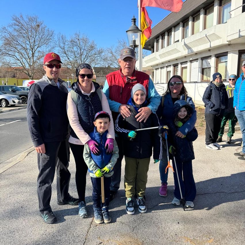 Eine Familie mit fünf Personen posiert für ein Foto vor einem Gebäude, hält Stöcke und Handschuhe. Im Hintergrund ist eine Fahne auf einem Pfahl gehisst.