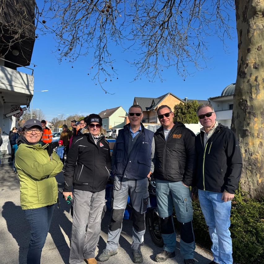 Fünf Personen, vier Männer und eine Frau, stehen in einer Reihe neben einem Baum und lächeln. Sie sind alle in Jacken und Hosen gekleidet, wobei einer der Männer eine Brille trägt. Hinter ihnen ist eine Menschenmenge zu sehen, und ein Gebäude mit Balkon befindet sich im Hintergrund.
