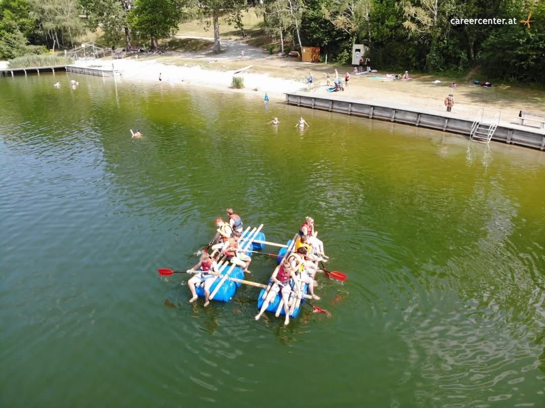 Eine Gruppe von Menschen paddelt auf zwei verbundenen Flößen in einem See, mit einem sandigen Strand und Bäumen im Hintergrund.
