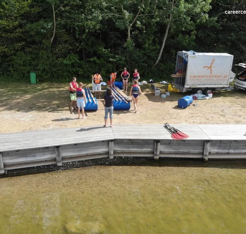 Eine Gruppe von Menschen in Schwimmwesten steht am Strand in der Nähe eines Holzstegs, mit einem LKW und Fässern in der Nähe.