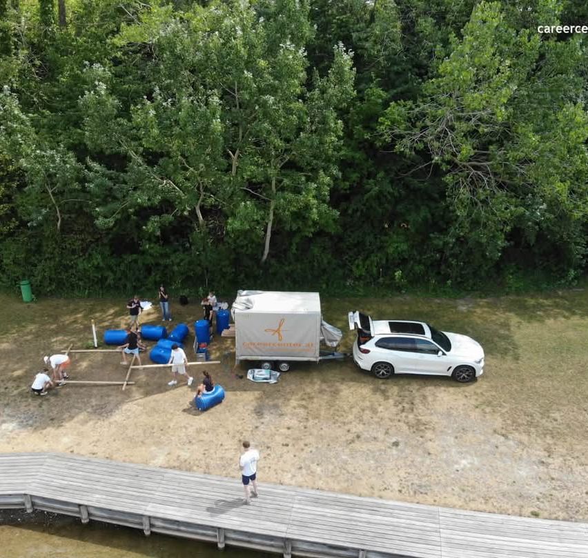 Eine Gruppe von Menschen arbeitet an einem Projekt in einem Waldgebiet. Ein weißer SUV ist in der Nähe eines Anhängers mit offenem Kofferraum geparkt. Einige Leute stehen herum, andere sitzen auf einem blauen Fass. Ein Holzweg führt zum Wasser.