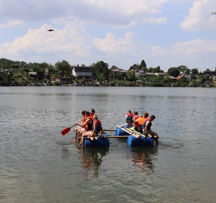 Eine Gruppe von Menschen in Schwimmwesten paddelt mit einem selbstgebauten Floß auf einem See, mit Häusern und Bäumen im Hintergrund.