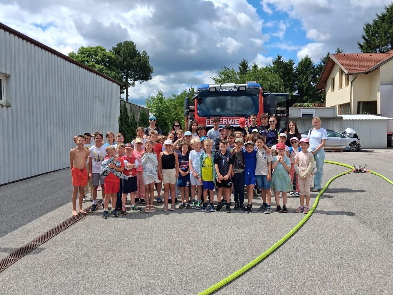 Eine Gruppe von Kindern und Erwachsenen steht vor einem roten Feuerwehrauto mit einer Schläuche auf dem Boden.