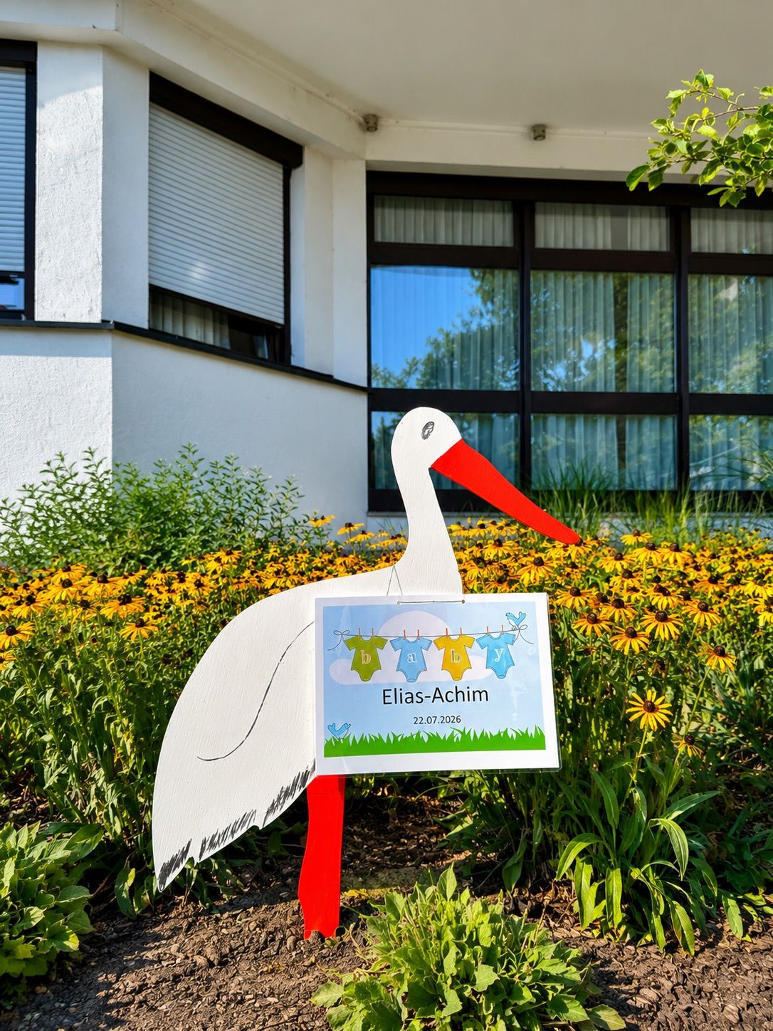 Ein Storch mit einem Schild, das Elias-Achim steht in einem Garten mit gelben Blumen. Dahinter befindet sich ein Gebäude mit Glasfenstern und einem Fensterrollo.
