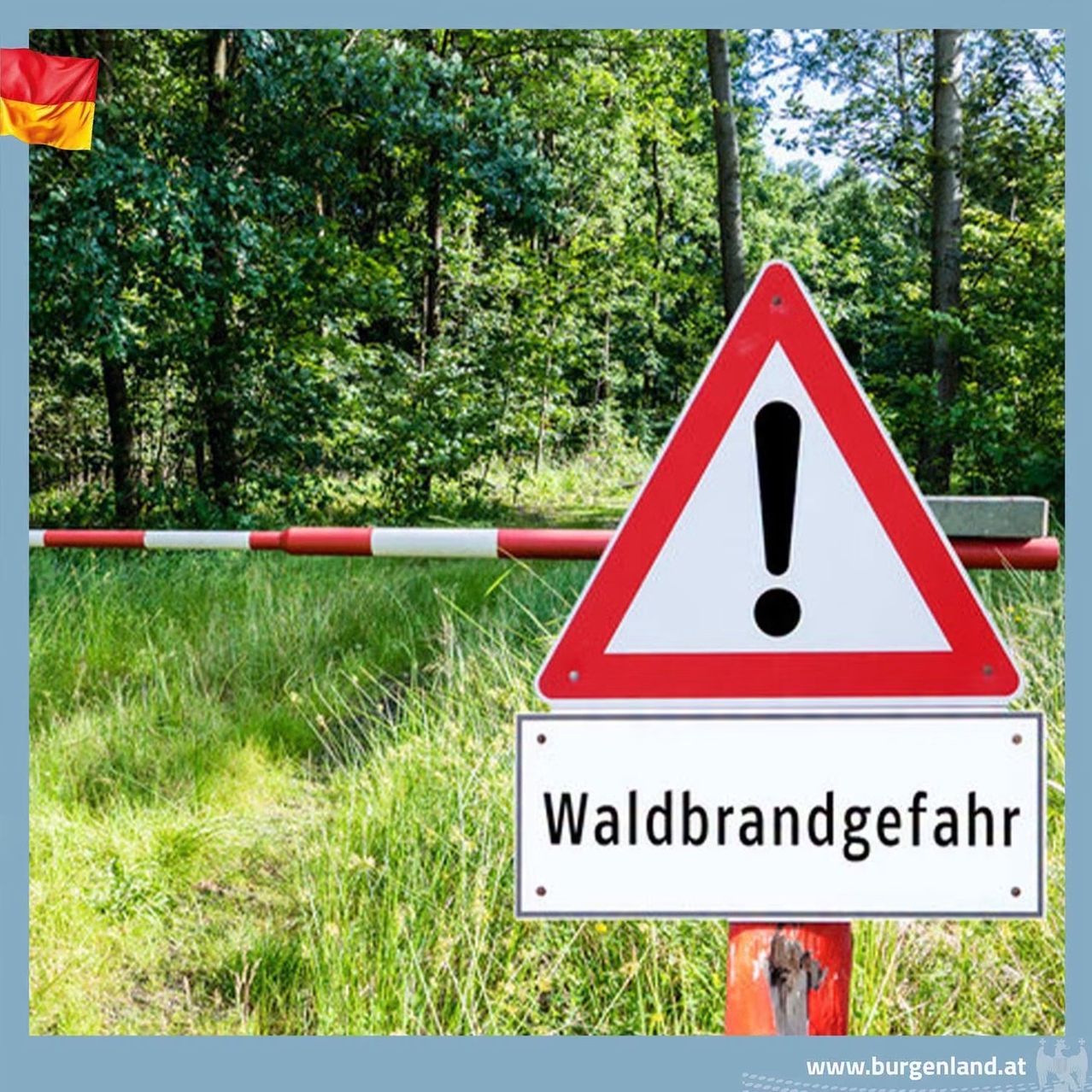 Ein rotes und weißes dreieckiges Warnschild mit Ausrufezeichen weist auf eine Waldbrandgefahr hin. Das Schild steht auf einem Pfosten in einer Grasfläche mit einer deutschen Flagge im Hintergrund.