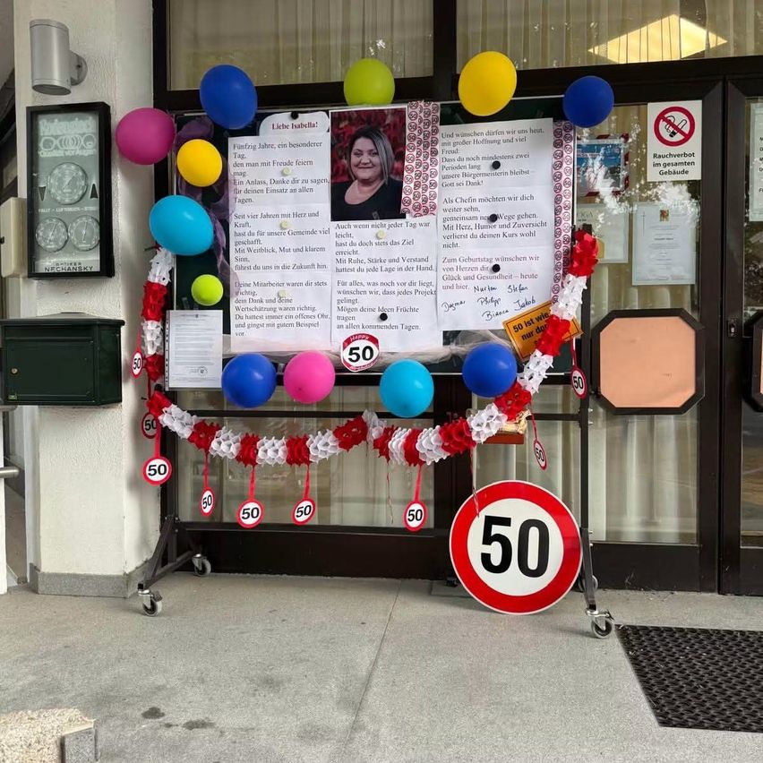 Eine Geburtstagsfeier vor einem Gebäude mit Luftballons, einem Schild und einem Foto einer Frau. Die Ballons sind bunt, und das Schild zeigt '50'. Es sind auch einige Papiere mit Schrift an den Ballons befestigt.