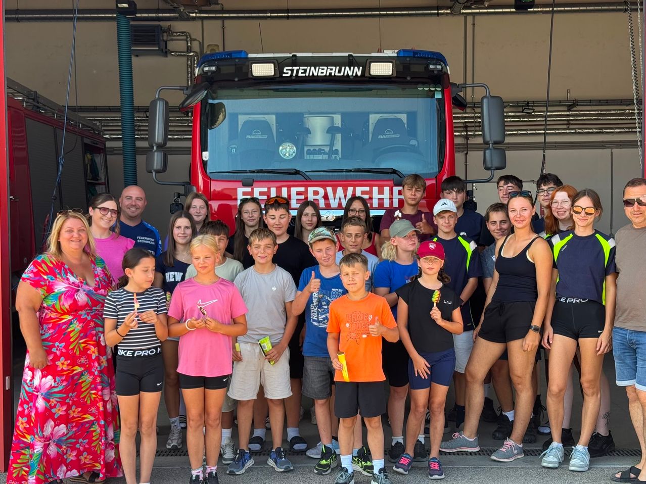 Eine Gruppe von Kindern und Erwachsenen posiert für ein Foto vor einem roten Feuerwehrauto mit der Aufschrift Steinbrunn. Sie halten Eislutscher und einige tragen Hüte.