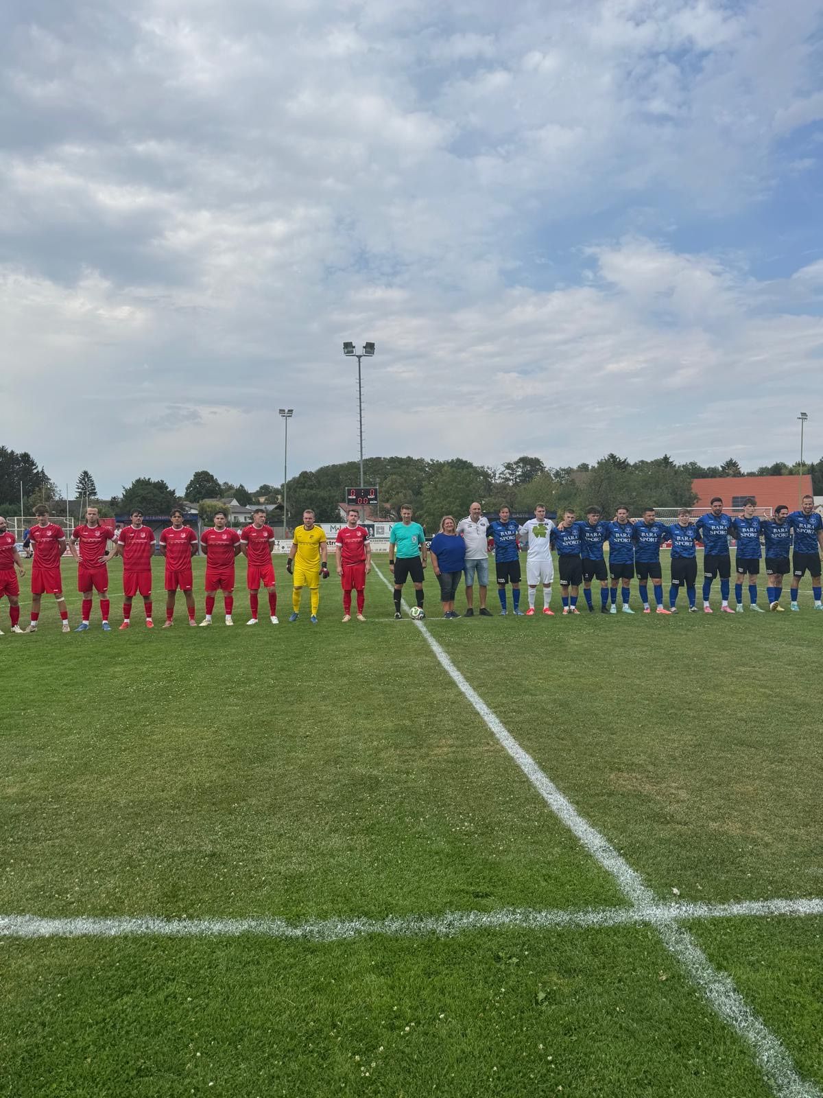 Two soccer teams line up on a field with a white line, a soccer ball, and a cloudy sky in the background.