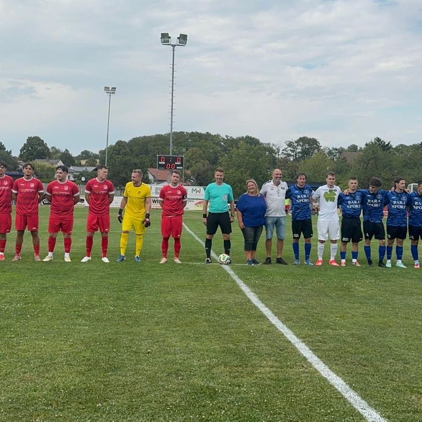 Two soccer teams stand on a field, one in red uniforms, the other in blue. The referee is in a green shirt, and a woman in a blue shirt stands among the players. Trees and a scoreboard are in the background.