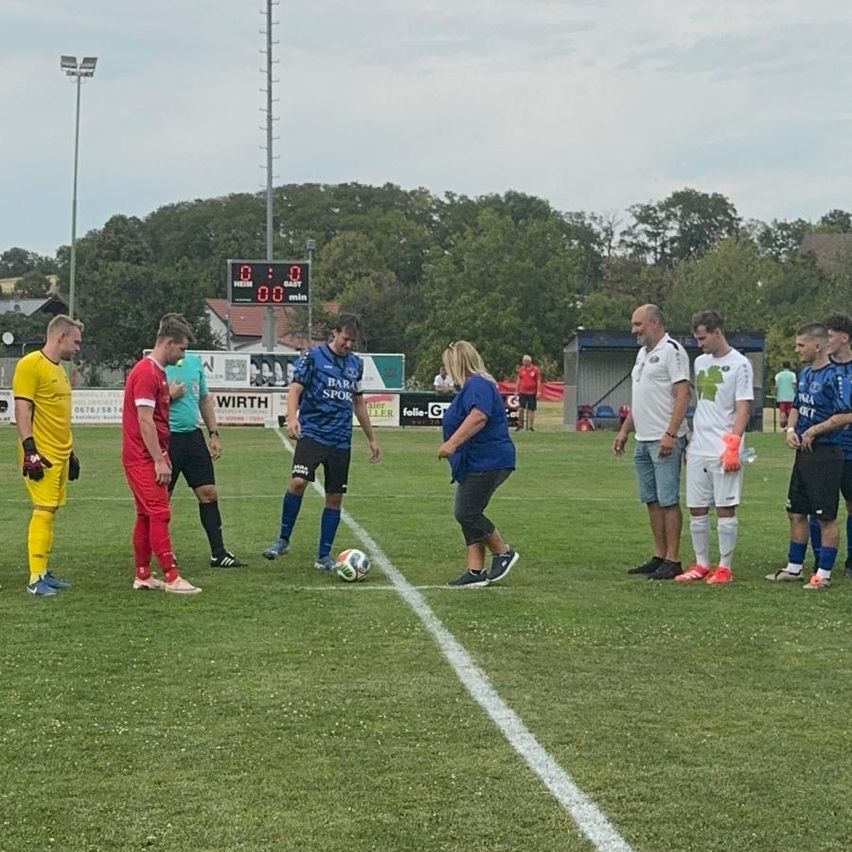 A soccer game is in progress on a field with players in different colored jerseys. A woman is standing near the ball, and several players are standing around, possibly waiting for the game to start. The scoreboard is visible in the background.
