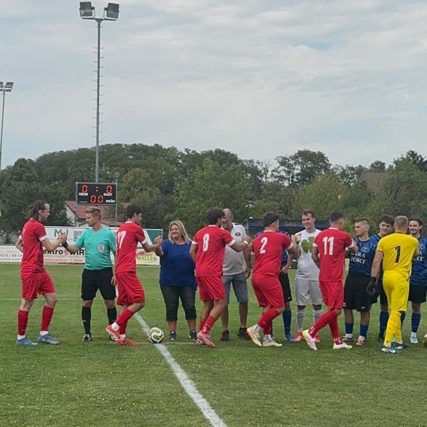 A group of soccer players in red uniforms, along with a referee and a woman, stand on a field with a soccer ball, under a cloudy sky.