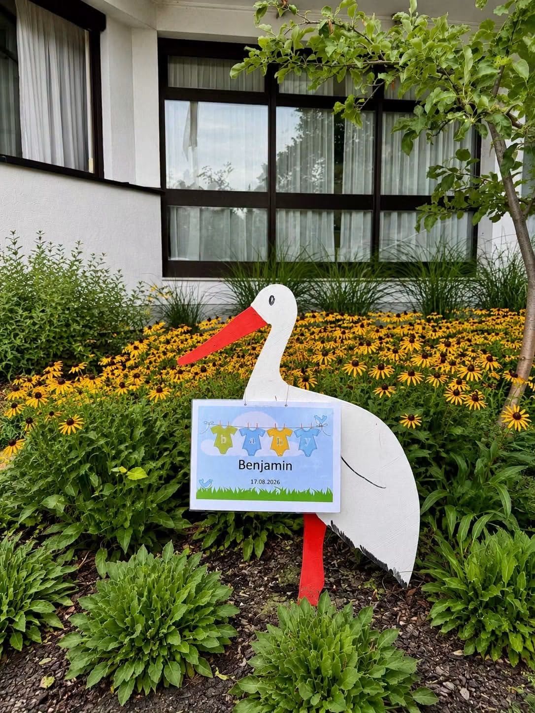 A stork statue stands in a garden with yellow flowers, holding a sign that reads 'Benjamin 17.08.2026'. Behind it, a building with windows and curtains is visible.