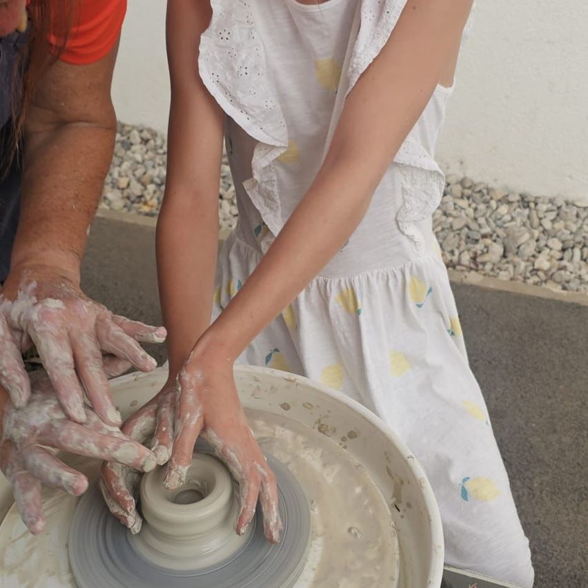 A young girl and an adult are working on a clay project on a pottery wheel. The girl is wearing a white dress with lemon patterns.