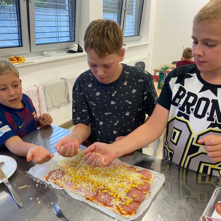 Three boys in a classroom are making a pizza with toppings like cheese and corn.