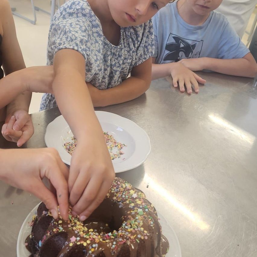 A young girl decorates a chocolate bundt cake with sprinkles at a table, with two other children watching.