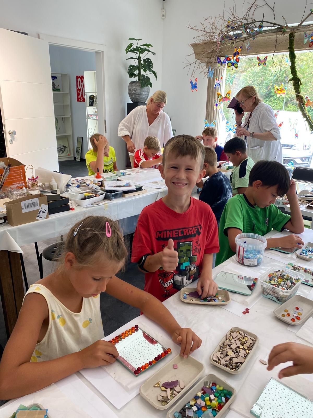 Children are engaged in a craft activity at a table, with a smiling boy giving a thumbs up. Various materials and tools are spread out on the table. An adult is supervising in the background.