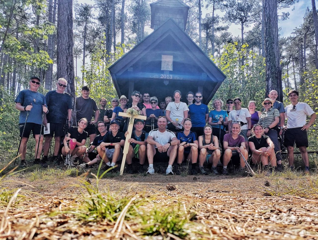 A group of people in outdoor gear pose for a photo in front of a small chapel with the year 2013 on its roof.