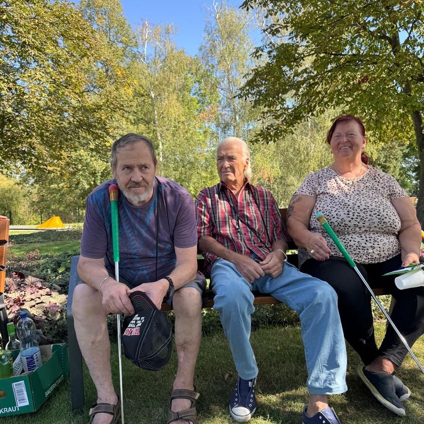 Three elderly individuals sit on a park bench, smiling. The man on the left holds a walking stick and a hat. The middle man wears a plaid shirt and blue jeans. The woman on the right holds a golf club.