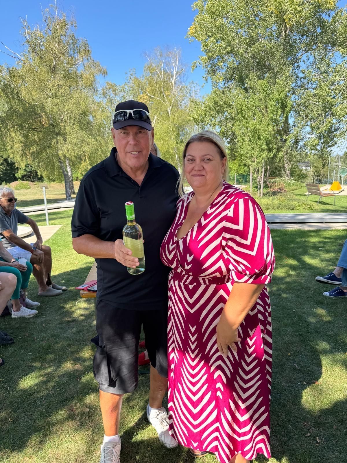 A man and a woman stand together in a park, the man holding a bottle of wine and smiling. Behind them, people sit on the grass, and trees and a bench are visible.