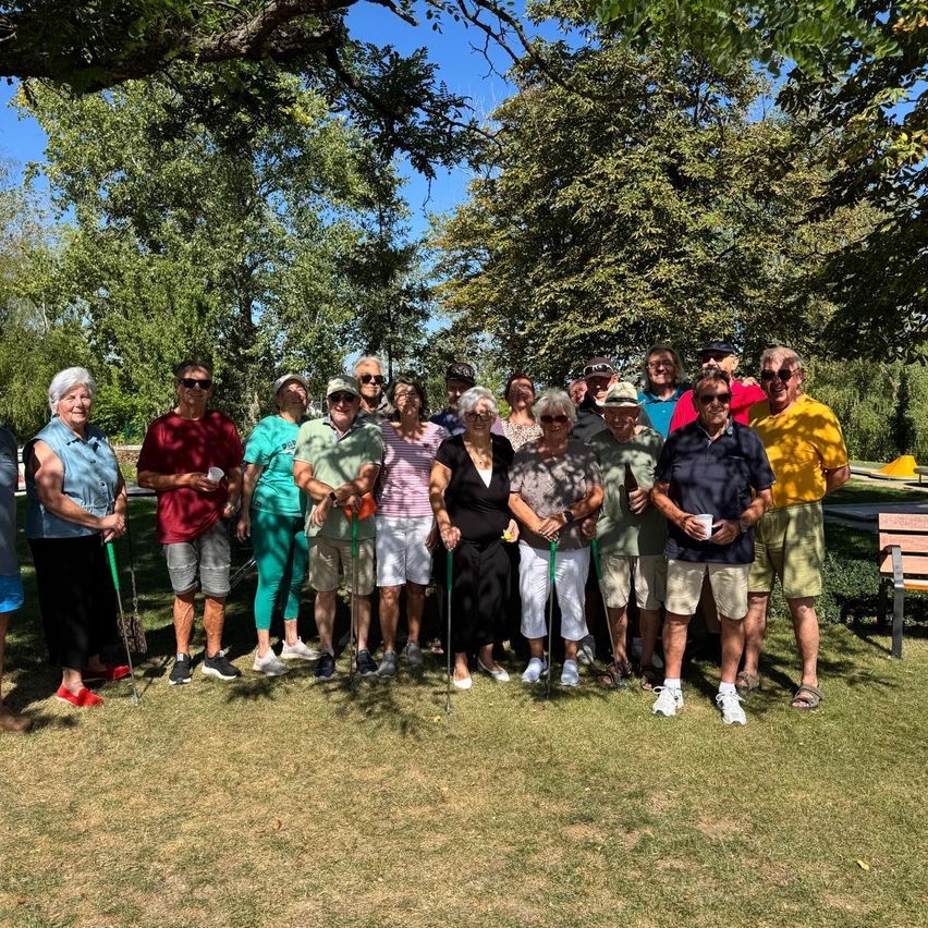 A group of people, possibly golfers, are posing for a photo in a park with trees and a bench in the background.
