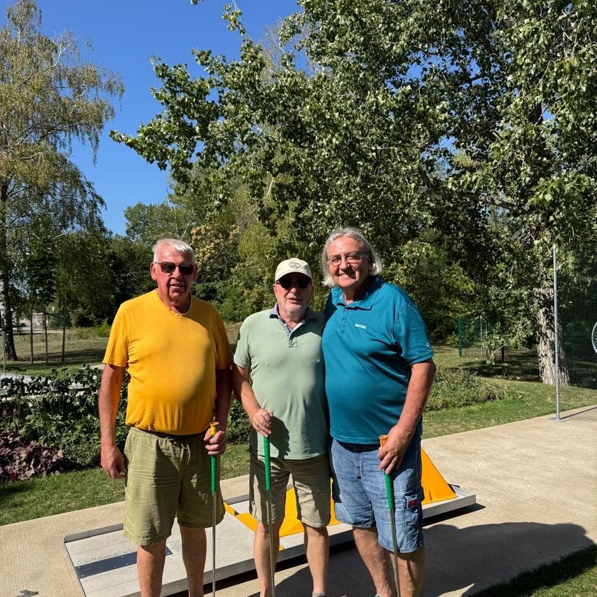 Three men stand on a golf course, smiling and holding golf clubs. The man on the left wears a yellow shirt, the middle one a green polo, and the right one a blue shirt. They are surrounded by trees and a fence.