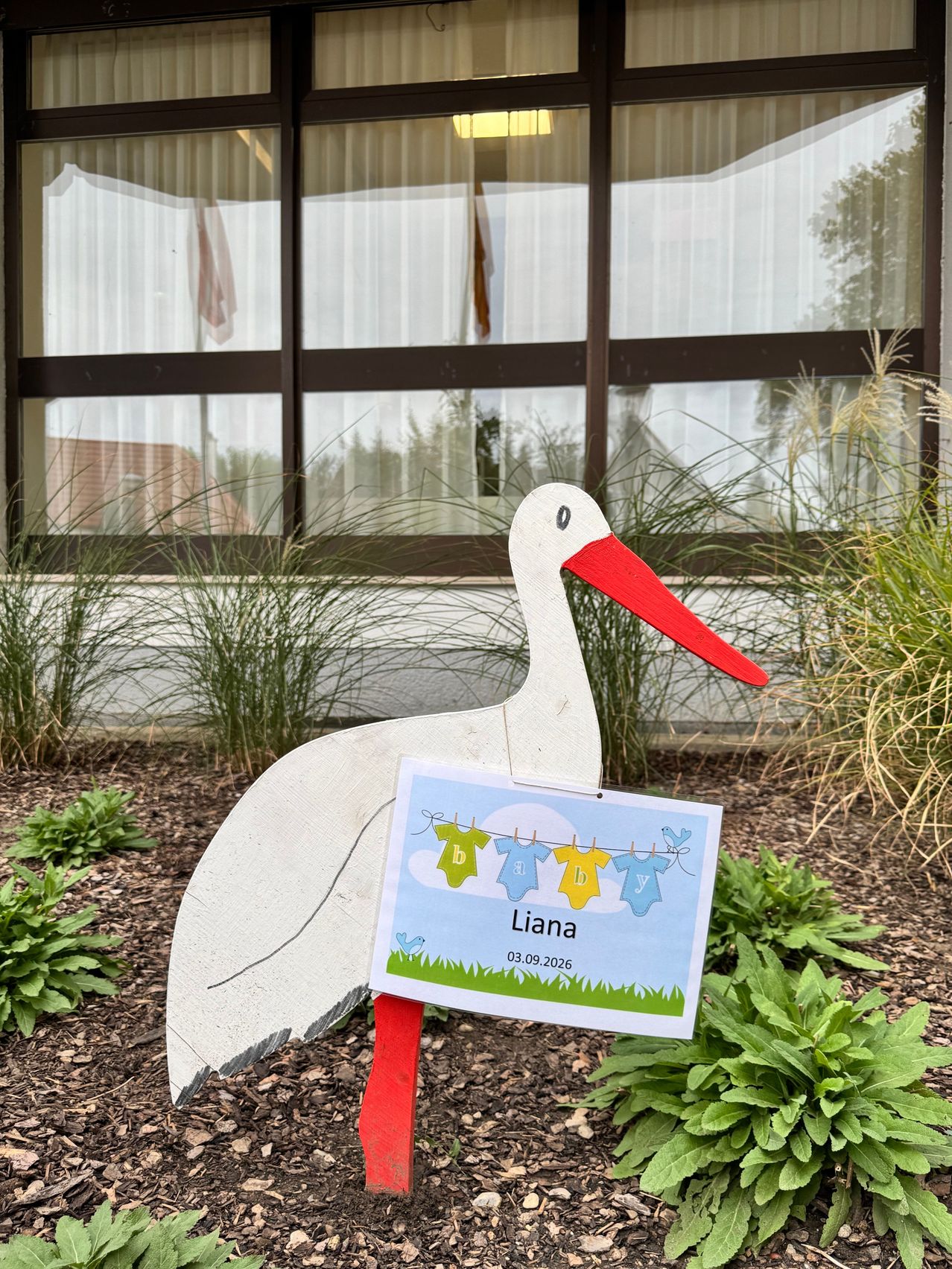 A stork statue with a sign reading 'Liana, 03.09.2026' stands in a garden with plants and a building in the background.