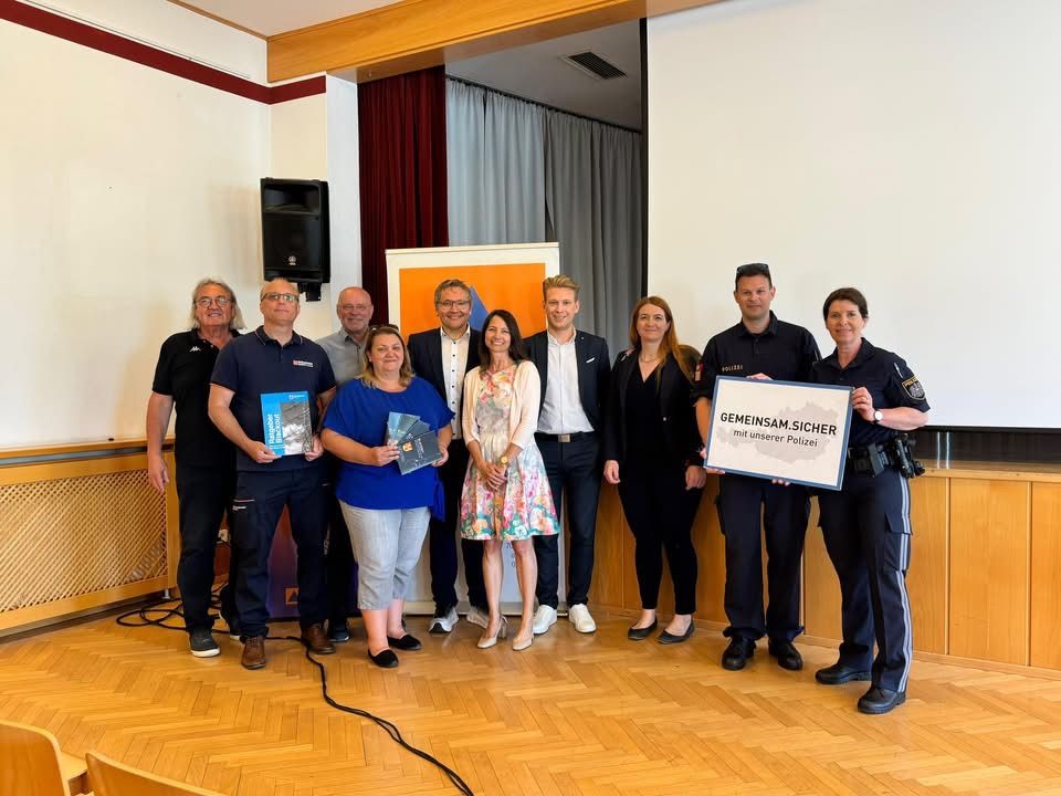 Eine Gruppe von Menschen steht in einem Raum, einige halten Bücher und ein großes weißes Schild. Eine Frau hält ein Schild mit der Aufschrift Gemeinsam.Sicher. mit unserer Polizei.
