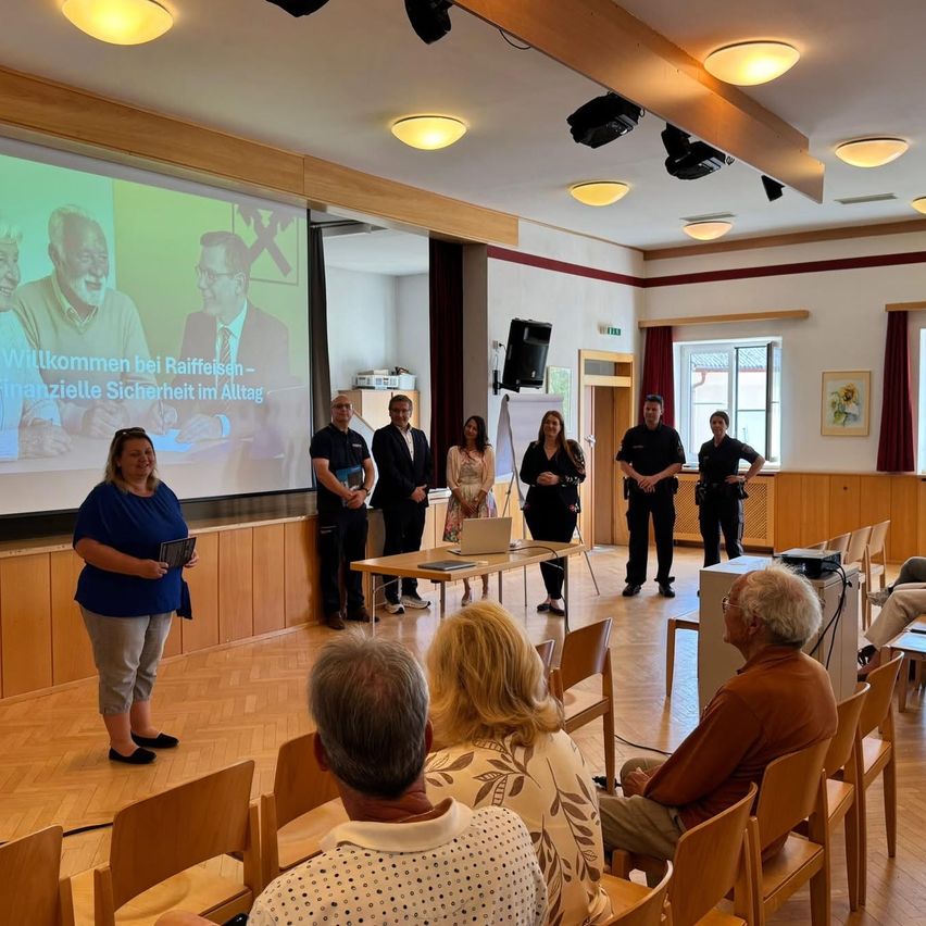 In einem Konferenzraum findet eine Präsentation über finanzielle Sicherheit statt. Zwei Männer und zwei Frauen stehen vorne, während mehrere Personen auf Stühlen sitzen.