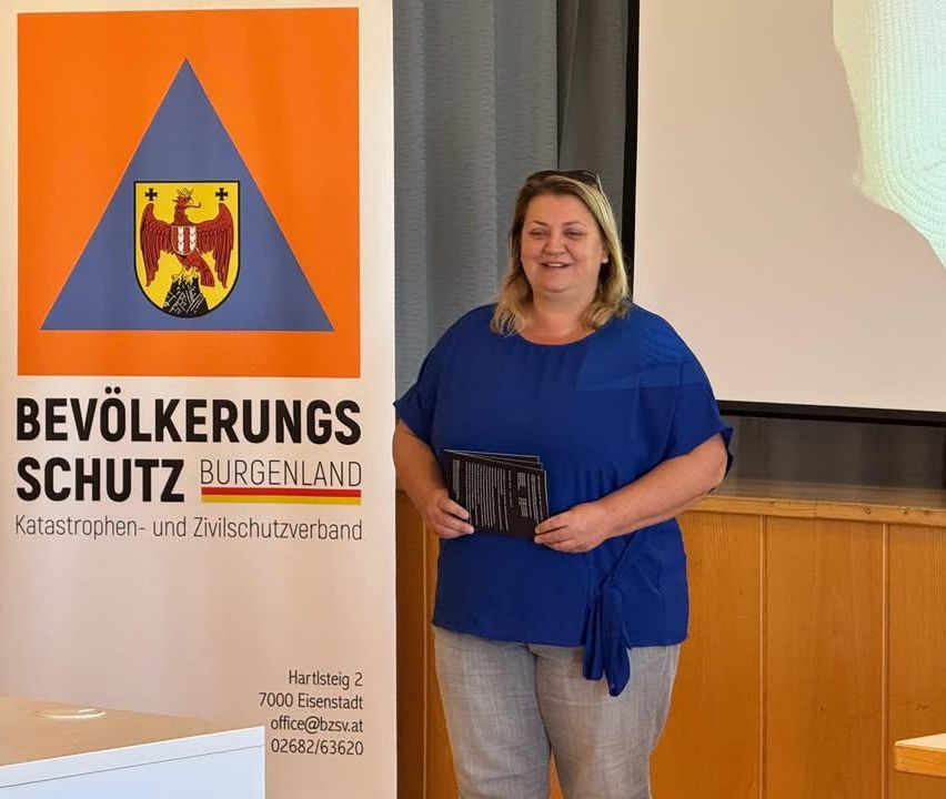 Eine Frau steht lächelnd vor einem Schild, das 'Evölkerungs Schutz Burgenland' mit einem Wappen und Kontaktdetails zeigt, während sie ein Büchlein in der Hand hält.