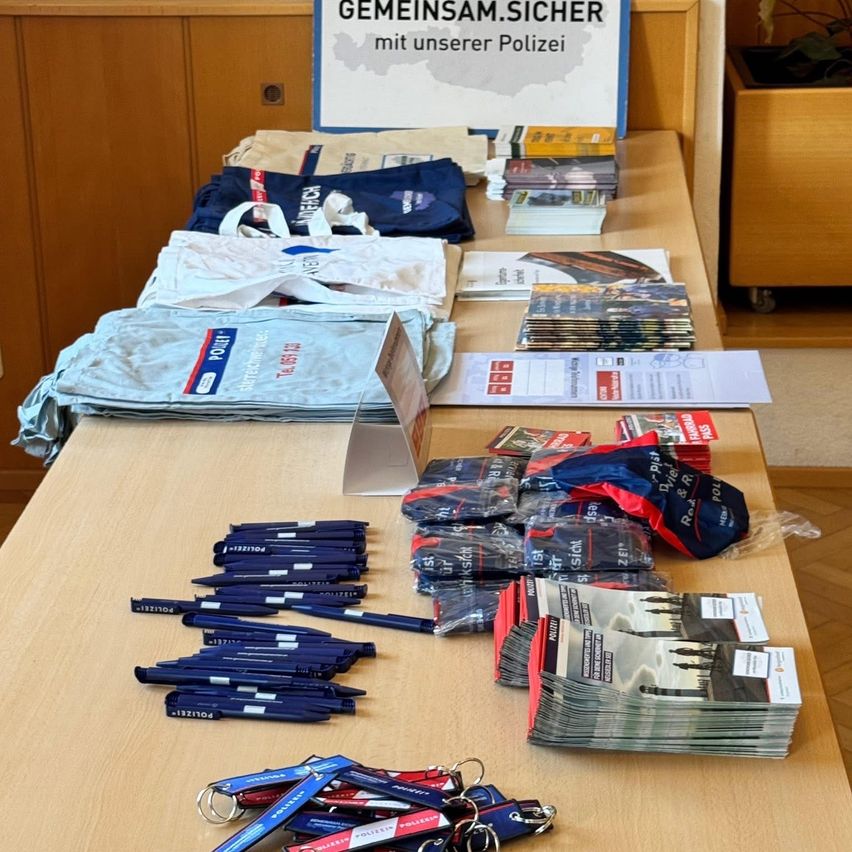 Ein Tisch ist mit Taschen, Stiften und Broschüren gefüllt. Ein Schild auf Deutsch steht 'Gemeinsam Sicher'.