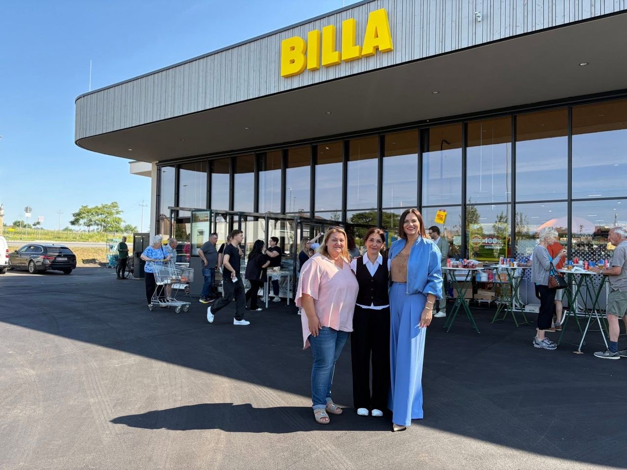 Drei Frauen stehen lächelnd vor dem Billa-Supermarkt. Dahinter gehen und schieben mehrere Personen Einkaufswagen. Der Laden hat große Fenster und ein gelbes Schild mit dem Wort Billa.